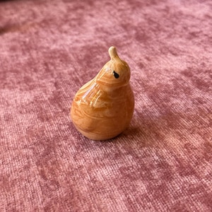 May include: A small, pear-shaped ceramic figurine with a light brown, wood-grain-like pattern. The figurine has a small, black dot for an eye and a small, white stem. The figurine is on a pink surface.