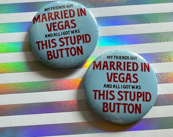 May include: Two light blue round buttons with the text "MY FRIENDS GOT MARRIED IN VEGAS AND ALL I GOT WAS THIS STUPID BUTTON" in red. The buttons are on a striped, iridescent background.