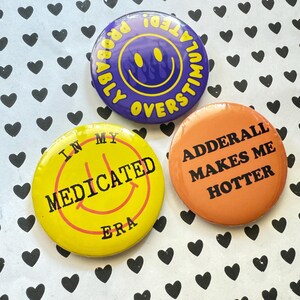May include: Three round buttons with text and smiley faces. One is purple with "Probably Overstimulated!" and a yellow smiley face. Another is yellow with "In My Medicated Era" and a smiley face. The last is orange with "Adderall Makes Me Hotter".