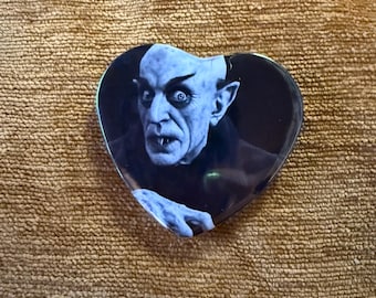 May include: Heart-shaped pin featuring a black and white image of a pale-skinned man with sharp teeth and intense eyes. The man is wearing a dark outfit. The pin is set against a textured, tan background.