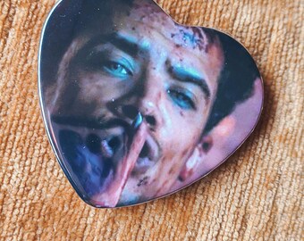 May include: A heart-shaped pin featuring a detailed photographic print of a person with bright blue eyes, dark hair, and a smudge on their face. The person is holding a thin, reddish-brown object near their mouth, with dark shadows and highlights.