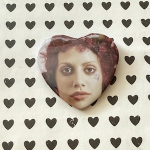 May include: Heart-shaped button featuring a portrait of a woman with red hair and a small square tattoo on her face. The button is set against a white background with a pattern of black hearts.