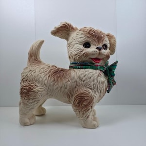 Puede incluir: Figura de perro vintage, crema con manchas marrones. Tiene una pajarita a cuadros verdes y rojos y una boca roja. El perro tiene ojos negros y está de pie. Un objeto decorativo encantador.
