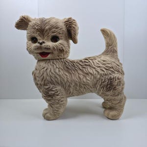 Puede incluir: Un perro de juguete vintage de color marrón claro con una apariencia texturizada y esponjosa. El perro tiene ojos marrones grandes, boca roja y orejas caídas. El juguete está de pie sobre sus cuatro patas, con la cola levantada.