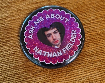 May include: A round button with a black border and a scalloped edge. The button features a photo of a man inside a heart-shaped cutout. The text "ASK ME ABOUT NATHAN FIELDER" is printed around the heart in white letters.
