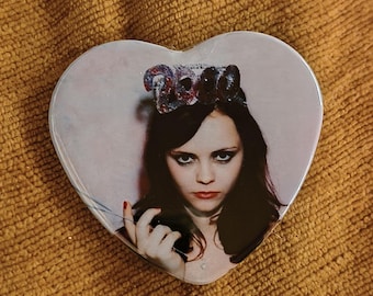 May include: A heart-shaped pin featuring a photographic print of a person with dark hair and dark eye makeup wearing a crown with the text 2000. The person has red lipstick and is holding a thin object near their face against a light pink background.