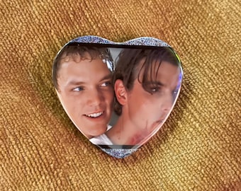 May include: A heart-shaped pin featuring a photographic print of two individuals. The person on the left has short, light brown hair and is smiling, while the person on the right has dark hair, dark eye makeup, and visible red marks on their neck.