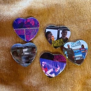 May include: Five heart-shaped pins with images from the movie "Dinner in America." Each pin features a different scene and the movie title. The pins have a glossy finish and are set against a golden-brown background.