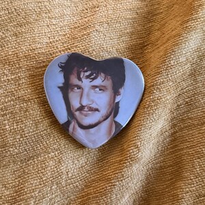 May include: A heart-shaped pin featuring a photographic portrait of a person with dark, wavy hair, a mustache, and a light beard. The image is rendered in muted, cool tones, showing the individual looking slightly to the side with a subtle smile.