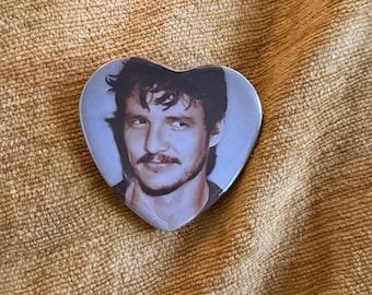 May include: A heart-shaped pin featuring a photographic portrait of a person with dark, wavy hair, a mustache, and a light beard. The image is rendered in muted, cool tones, showing the individual looking slightly to the side with a subtle smile.