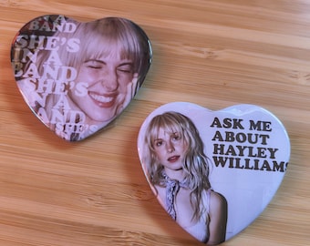 May include: Two heart-shaped pin-back buttons featuring photographs of Hayley Williams. The left button displays the text "A BAND SHE'S IN A BAND SHE'S IN A BAND SHE'S". The right button displays the text "ASK ME ABOUT HAYLEY WILLIAMS".