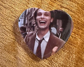 May include: Heart-shaped button featuring a smiling person in a brown suit and tie. The image is a photograph of a person in a city setting. The button is set against a textured, yellow-orange background.