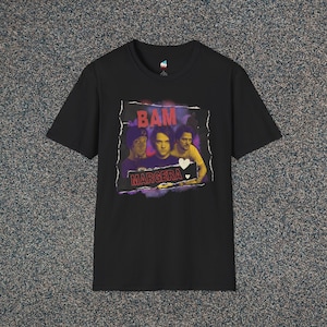 May include: Black t-shirt featuring a graphic of three people with the text "BAM MARGERA" in a distressed style. The graphic has purple, red, and yellow accents. The shirt is made of a soft material.