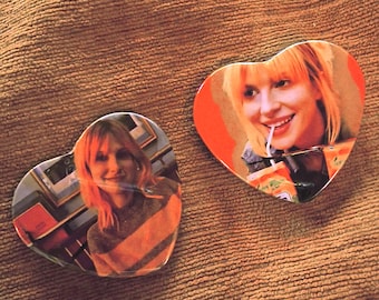 May include: Two heart-shaped coasters featuring a woman with blonde hair. One coaster has a photo of the woman in a striped sweater, and the other shows her drinking from a cup with a straw. The coasters are on a brown surface.