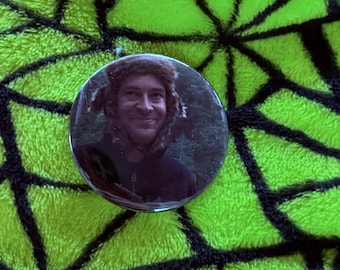 Peut inclure: Un bouton rond représentant une personne souriante portant une capuche bordée de fourrure. Le bouton est posé sur un fond vert vif texturé avec un motif en forme de toile d'araignée noire.