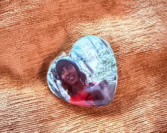 May include: A heart-shaped clear acrylic charm with a photograph of a person wearing a hat and a red shirt. The charm is resting on a textured, orange-brown surface.