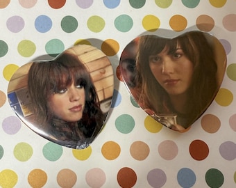 May include: Two heart-shaped pins featuring photographic portraits of a person with long brown hair and bangs. The left pin shows the person looking to the side, while the right pin shows the person looking forward, holding a small silver camera.