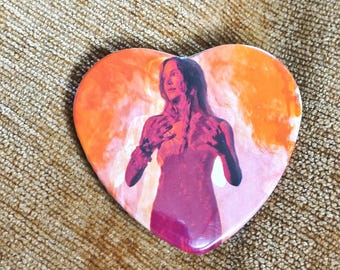 May include: A heart-shaped pin featuring a graphic illustration of a person with long hair, positioned centrally against a vibrant background of swirling orange and pink smoke. The figure is depicted with hands raised near the chest area.