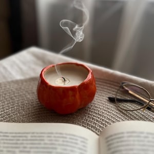 Könnte beinhalten: Ein kleiner, orangefarbener, kürbisförmiger Kerzenhalter mit einer brennenden Docht und Rauchschwaden. Die Kerze steht auf einer strukturierten Oberfläche neben einem offenen Buch und einer Brille, was eine gemütliche, herbstliche Szene schafft.