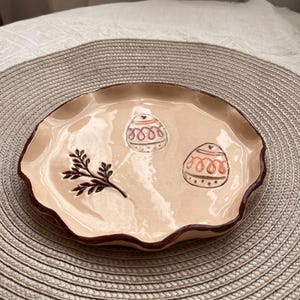 May include: A beige ceramic plate with a wavy edge and a dark brown rim. The plate features two decorated egg designs and a sprig of leaves. The plate is sitting on a woven placemat.