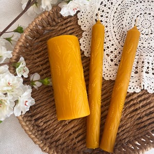 May include: Three yellow beeswax candles and one pillar candle with wheat stalk designs, arranged in a woven basket. The candles are on a white lace doily, with white flowers in the background.