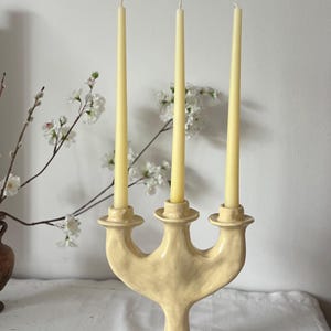 May include: A pale yellow ceramic candelabra with a textured, organic surface featuring three distinct candle holders. Three slender, light cream-colored taper candles are inserted into the holders, standing upright in a symmetrical arrangement.
