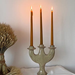 May include: A three-armed ceramic candelabra with a glossy, light-toned glaze, holding three lit, slender, golden-brown taper candles. The candelabra features a curved, symmetrical base and three distinct candle cups, each supporting a burning candle.