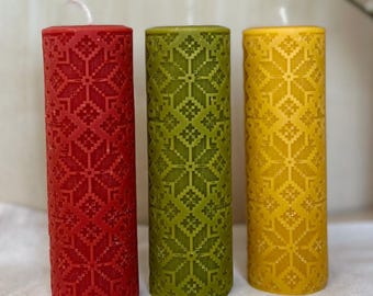 May include: Three pillar candles in red, green, and yellow. Each candle has a repeating geometric pattern carved into the wax. The candles are cylindrical and stand upright on a white surface. The candles are unscented.