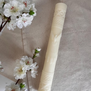 Puede incluir: Una vela cilíndrica alta de color blanco roto con patrones decorativos en relieve, que incluyen un ave central con las alas extendidas, entramados geométricos en la parte superior e inferior y espigas de trigo verticales.