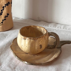 Könnte beinhalten: Eine hellgelbe Keramiktasse in Kürbisform mit passender Untertasse. Die Tasse hat einen dicken Henkel und eine gesprenkelte Glasur. Die Untertasse besitzt einen gewellten Rand und einen griffartigen Fortsatz an einer der Seiten.
