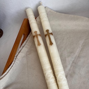 Op de afbeelding: Twee ivoorkleurige kaarsen met een textuurpatroon en gouden kruisen. De cilindrische kaarsen hebben een decoratief ontwerp. Ze liggen op een houten dienblad met een neutrale doek. De kaarsen zijn ongeveer 20 cm lang.