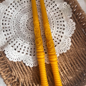 Puede incluir: Dos velas amarillas de cera de abeja con intrincados diseños en relieve, incluyendo cruces y patrones decorativos. Las velas tienen una base acanalada y se exhiben sobre un mantel de encaje blanco dentro de una cesta tejida.