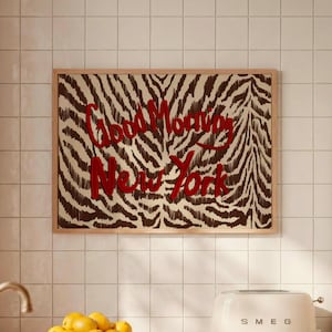 Könnte beinhalten: Ein rechteckiger Wandkunstdruck in einem hellen Holzrahmen mit einem braun-cremefarbenen Zebramuster und dem roten Text Good Morning New York, der mittig auf der Oberfläche platziert ist. Das Kunstwerk hängt an einer weiß gefliesten Küchenwand.