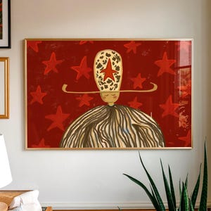 May include: A framed art print with a whimsical illustration. The artwork shows a figure with a striped body and a patterned hat with a red star. The background is a deep red with scattered red stars. The frame is gold.
