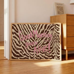 Op de afbeelding: Een ingelijst kunstwerk met een zebraprint achtergrond en de roze tekst "Good Morning you're Alive!". Het kunstwerk zit in een licht houten frame en staat op een houten vloer.