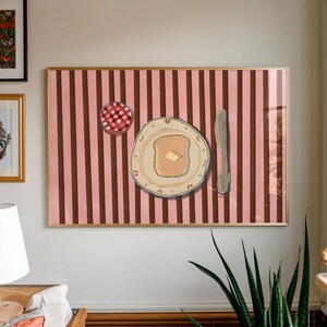 Puede incluir: Obra de arte enmarcada que representa una escena de desayuno. Un plato con tostadas y mantequilla, un cuchillo y un tarro de mermelada se representan sobre un fondo a rayas rosas y marrones. La obra de arte est&aacute; en un marco dorado y cuelga de una pared blanca.