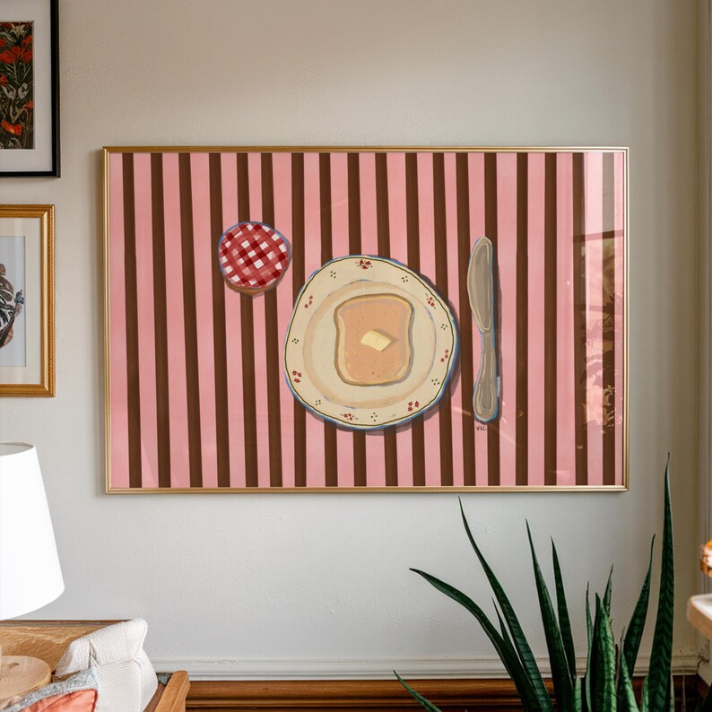 Puede incluir: Obra de arte enmarcada que representa una escena de desayuno. Un plato con tostadas y mantequilla, un cuchillo y un tarro de mermelada se representan sobre un fondo a rayas rosas y marrones. La obra de arte est&aacute; en un marco dorado y cuelga de una pared blanca.