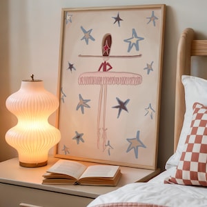 May include: A framed artwork featuring a ballerina with a star-shaped headpiece and a tutu, surrounded by blue and gold stars. A glowing, ribbed glass lamp and an open book sit on a nightstand. A bed with a patterned pillow and pink blanket is in the foreground.