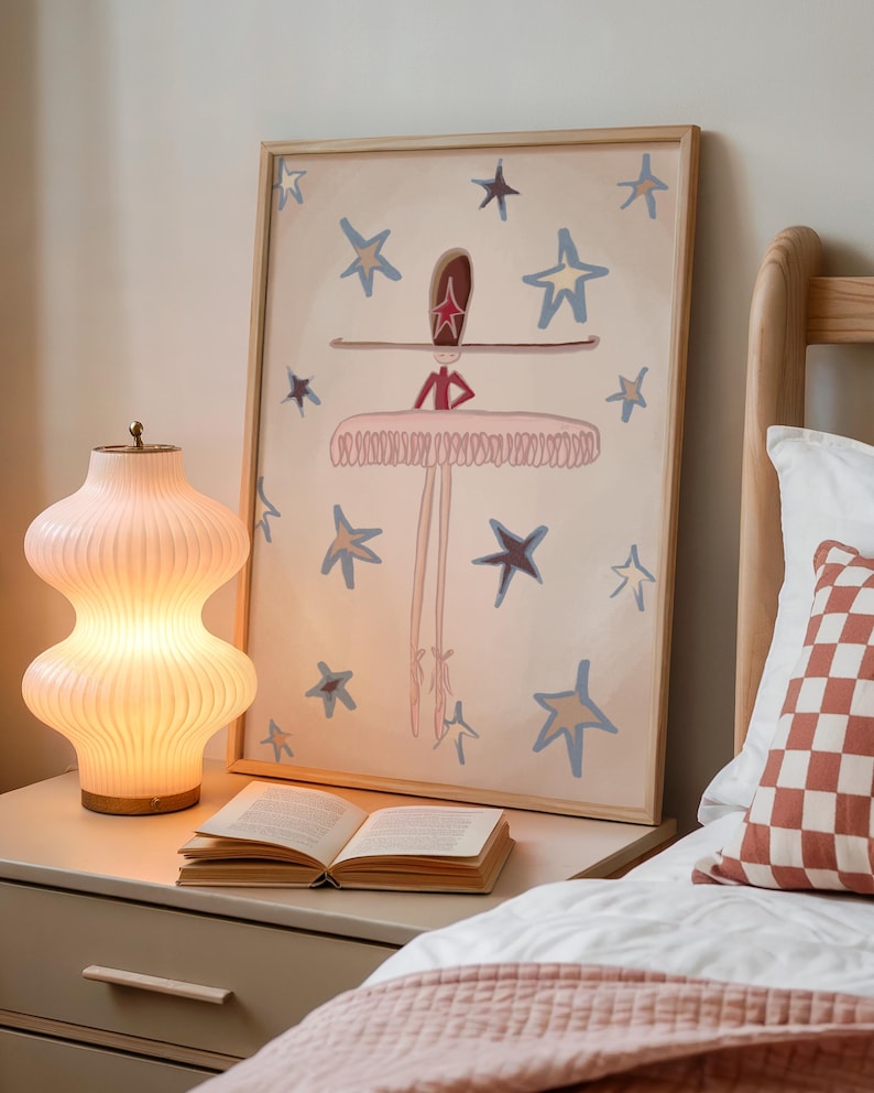 May include: A framed artwork featuring a ballerina with a star-shaped headpiece and a tutu, surrounded by blue and gold stars. A glowing, ribbed glass lamp and an open book sit on a nightstand. A bed with a patterned pillow and pink blanket is in the foreground.