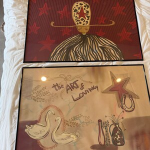 May include: Two framed art prints. The top print features a figure with a star-shaped head and a striped dress against a red star-filled background. The bottom print has swans, a star, a horseshoe, and the text "the ART of LOVING".