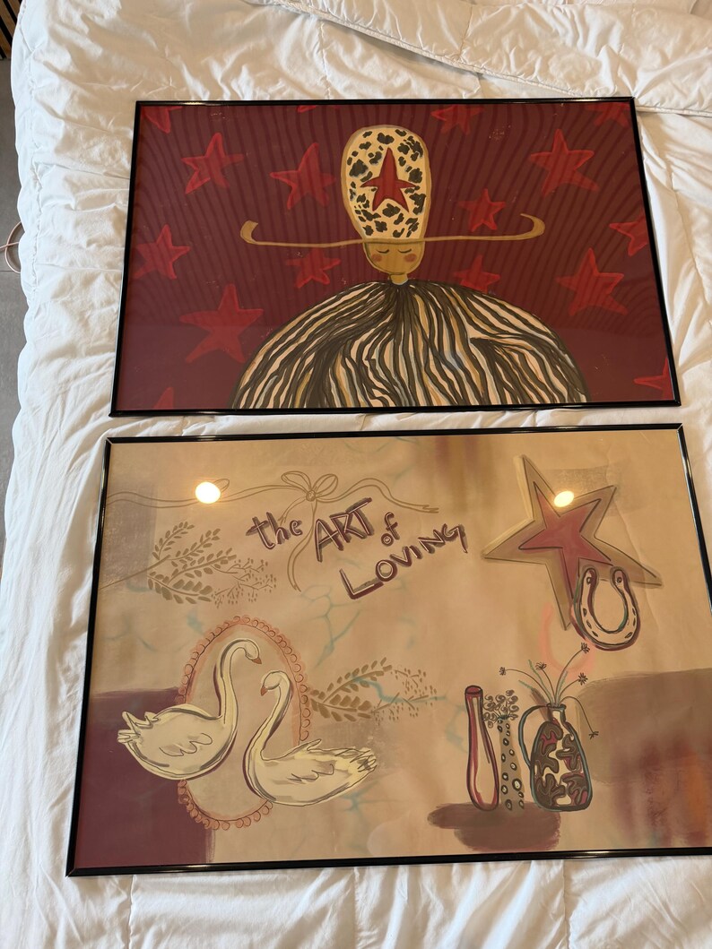 May include: Two framed art prints. The top print features a figure with a star-shaped head and a striped dress against a red star-filled background. The bottom print has swans, a star, a horseshoe, and the text "the ART of LOVING".
