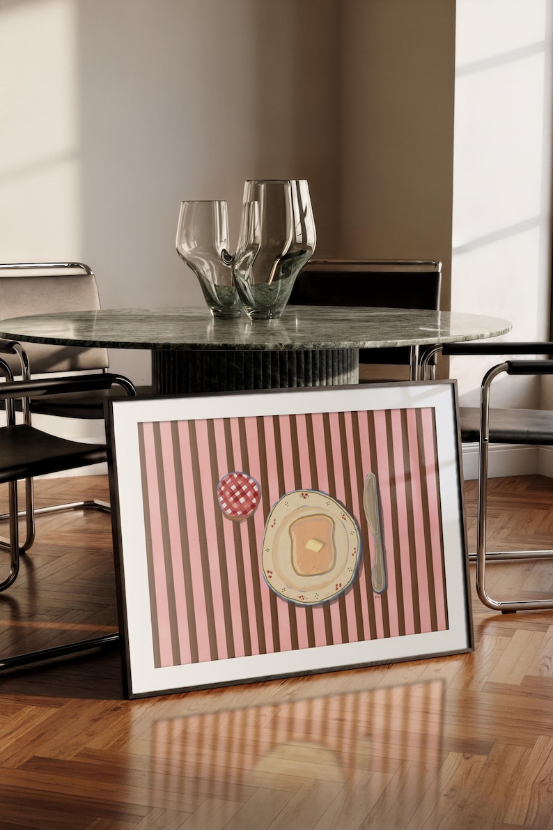 Puede incluir: Impresi&oacute;n art&iacute;stica enmarcada que representa un desayuno: tostadas con mantequilla, mermelada y un cuchillo, sobre un fondo a rayas rosas y marrones. Dos jarrones de cristal decorativos sobre una mesa de m&aacute;rmol. Marco negro.