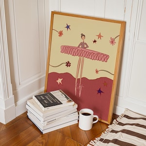 May include: A framed illustration of a ballerina in a pink tutu, with a light yellow background and a burgundy base. The artwork is propped against a white wall, next to a stack of books and a white coffee mug.