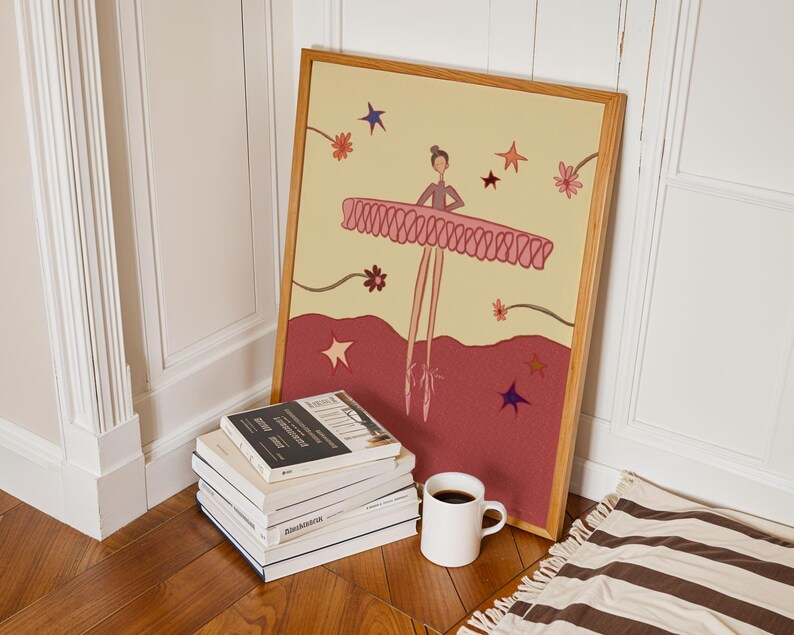 May include: A framed illustration of a ballerina in a pink tutu, with a light yellow background and a burgundy base. The artwork is propped against a white wall, next to a stack of books and a white coffee mug.