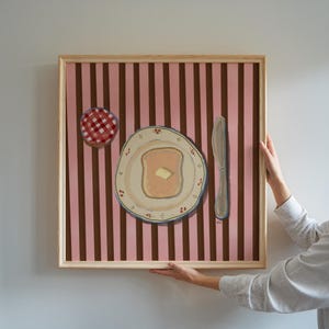 Puede incluir: Pintura enmarcada que representa una escena de desayuno. La obra muestra una rebanada de pan tostado con mantequilla en un plato decorativo, un tarro de mermelada y un cuchillo sobre un fondo a rayas rosas y marrones. El marco es de madera clara.