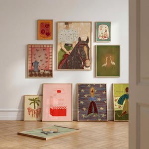May include: A collection of framed wall art prints featuring various illustrations including a person on a horse, a moka pot, cranes, a carton labeled Creative Juice, and abstract figures, displayed on a wall and on a wooden floor.