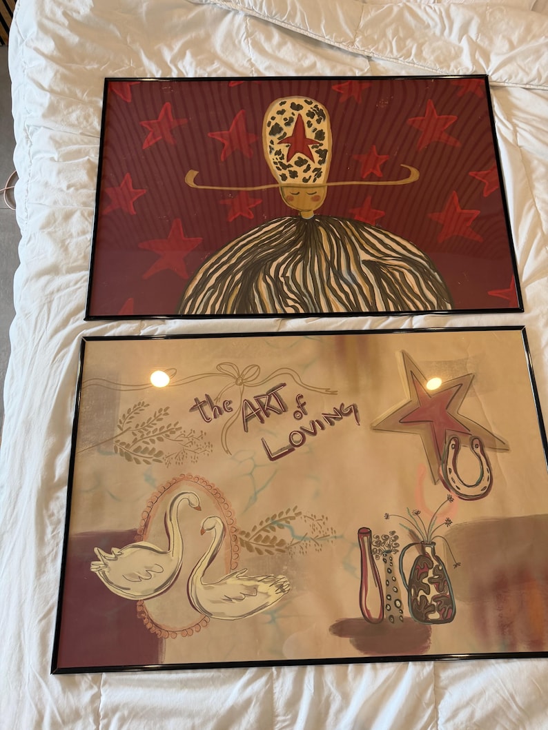 May include: Two framed art prints. The top print features a figure with a star-shaped head and a striped dress against a red star-filled background. The bottom print has swans, a horseshoe, and the text "the ART of LOVING".