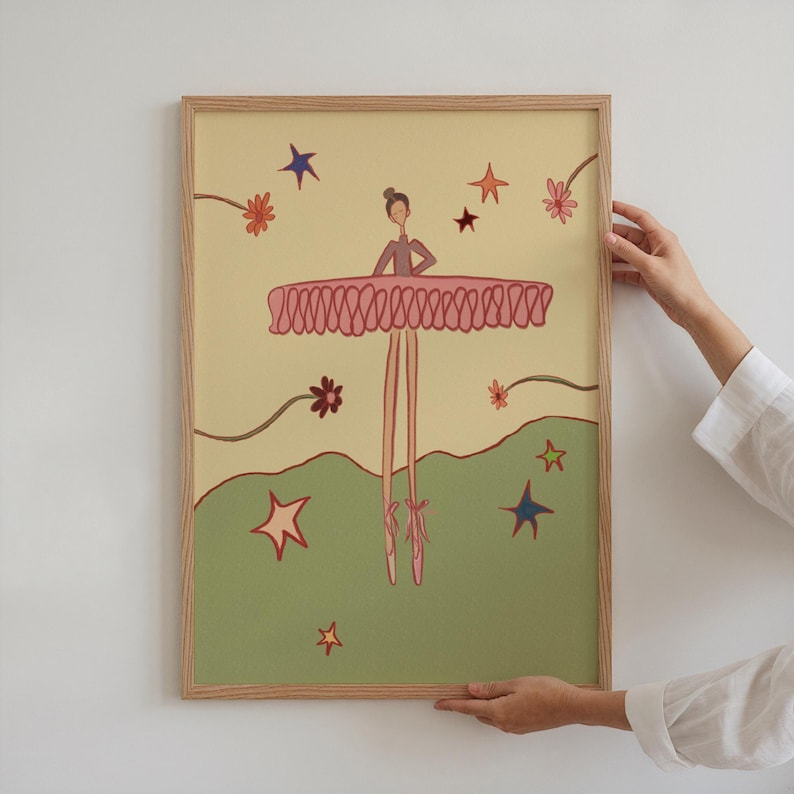May include: A framed illustration of a ballerina in a pink tutu and pointe shoes, with a light yellow background and green ground. The artwork features stars and flowers in various colors, with a light brown wooden frame.