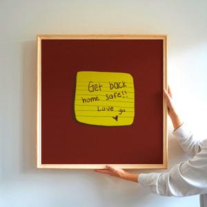 Pode incluir: Um quadro de avisos emoldurado com um fundo bordô e um post-it amarelo. A nota diz "Get back home safe!! Love ya" em tinta preta. A moldura é feita de madeira clara.