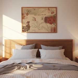 Puede incluir: Una escena de dormitorio con un cabecero de madera y una cama con ropa de cama a rayas y almohadas grises. Encima de la cama cuelga una obra de arte enmarcada con ilustraciones de una estrella, cerezas, flores y cisnes. La obra de arte incluye el texto "HOW LUCKY ARE WE".